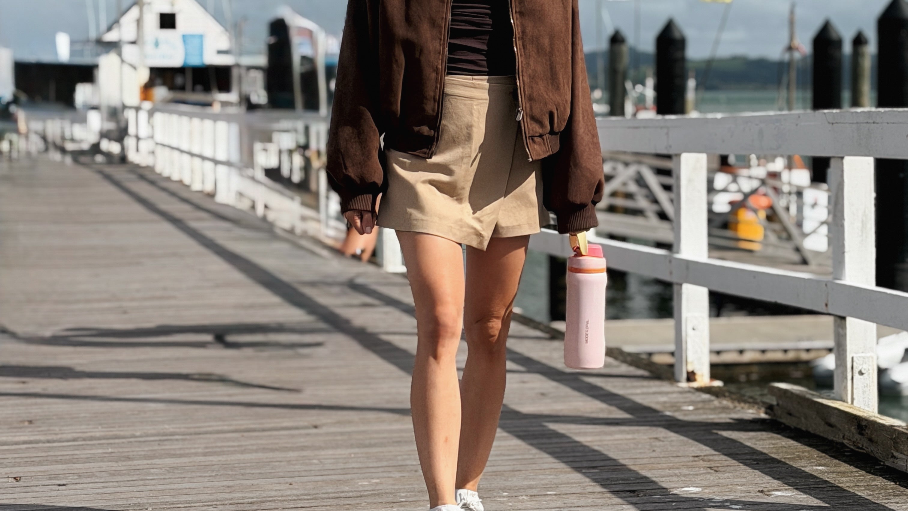 A woman walks on a wooden pier wearing a brown bomber jacket, beige shorts, a dark top, and white sneakers, holding a pink water bottle under her arm.