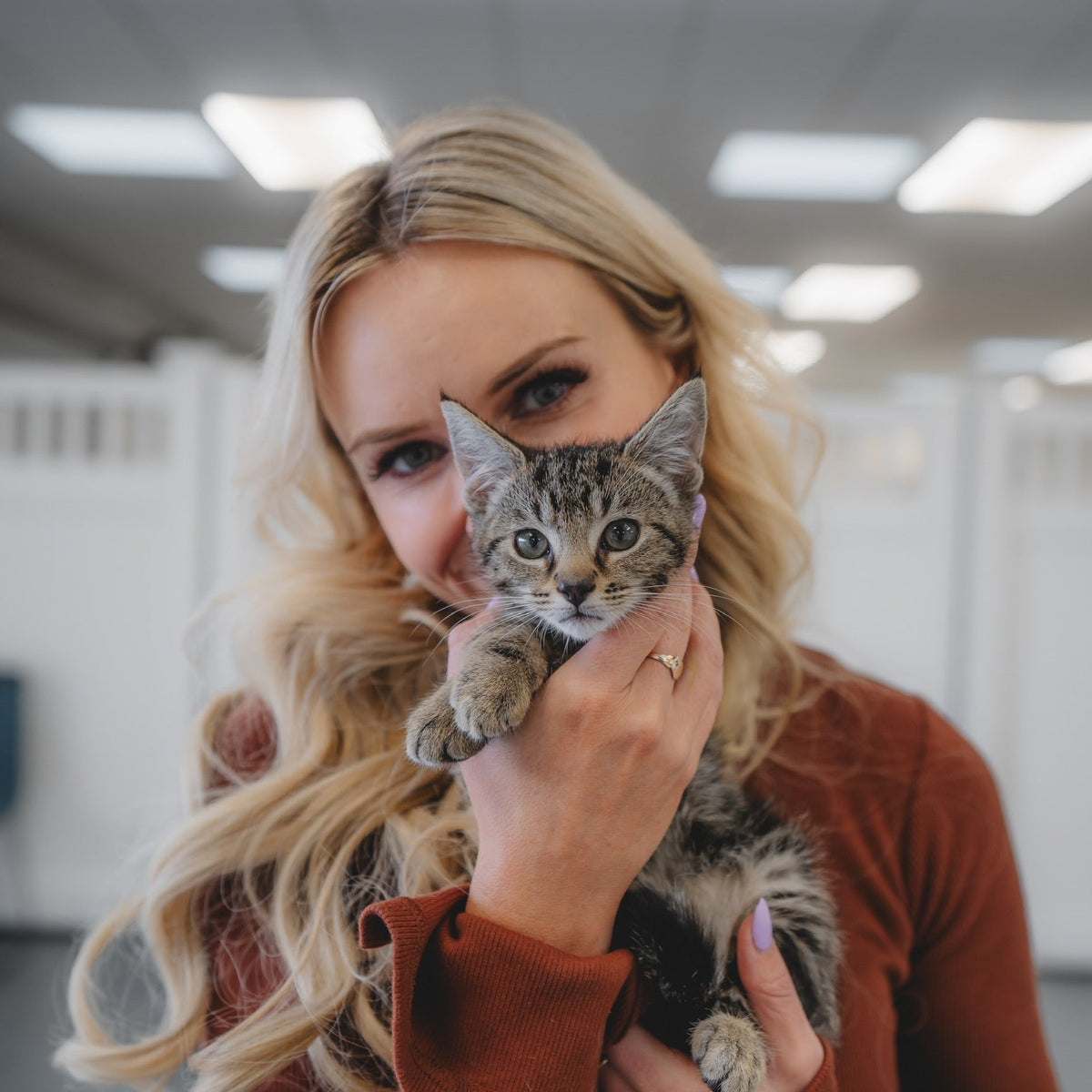 A woman with long blonde hair holds a small tabby kitten close to her face. She is wearing a rust-brown long-sleeve top, and the background shows a softly blurred indoor space with ceiling lights.